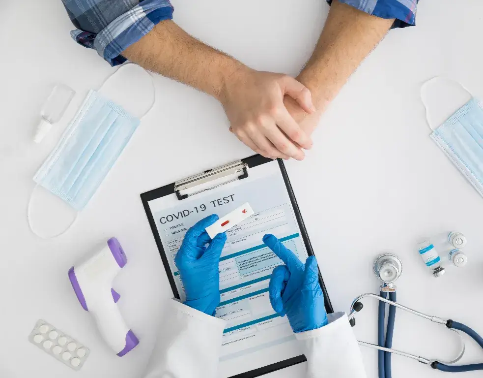 Doctor in blue gloves holding a COVID-19 test kit and pointing at a test form, with medical supplies and a stethoscope on a table, emphasizing at-home lab testing services and health management.